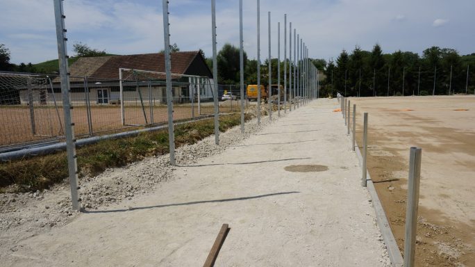 Ein unvollendeter Weg neben einem Sportfeld mit Zaun und angrenzendem Gebäude.
