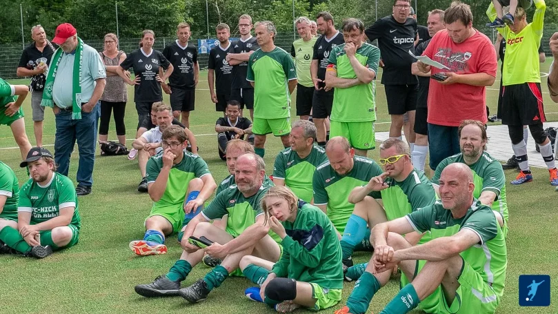 Eine Gruppe von Fußballspielern in grünen Trikots sitzt auf dem Rasen, Zuschauer im Hintergrund.