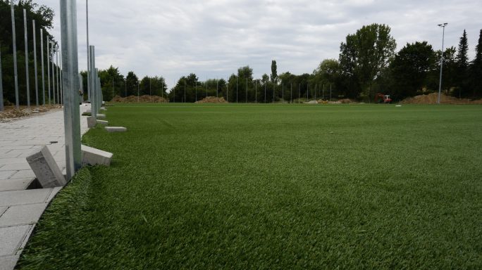 Fußballfeld mit grünem Kunstrasen und bewaldetem Hintergrund. Wolkiger Himmel.