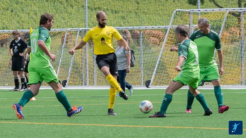 Zwei Mannschaften in grünen Trikots spielen Fußball auf einem Platz; ein Schiedsrichter leitet das Spiel.