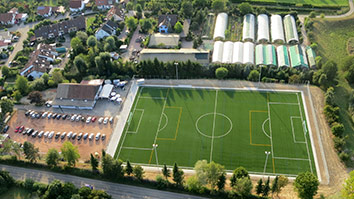 Luftaufnahme eines Fußballfeldes mit angrenzenden Parkplätzen und Wohngebieten.