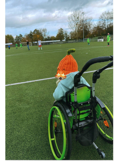 Ein Kind im Rollstuhl mit einem orangefarbenen Hut schaut ein Fußballspiel.