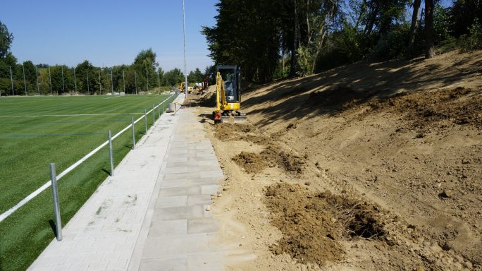 Baustelle am Sportplatz mit erdigem Hang und Maschinen neben einem Gehweg.