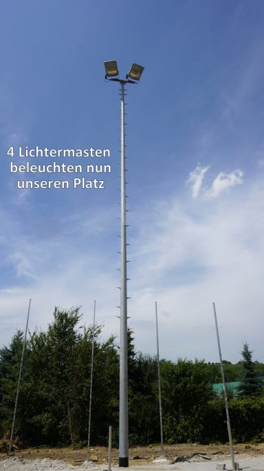 Ein hoher Lichtmast mit vier Leuchten vor einem blauen Himmel und Bäumen.