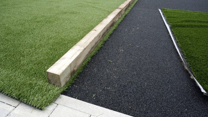 Grüner Rasen trifft auf schwarzen Asphalt, mit Holzrand zwischen den Flächen.