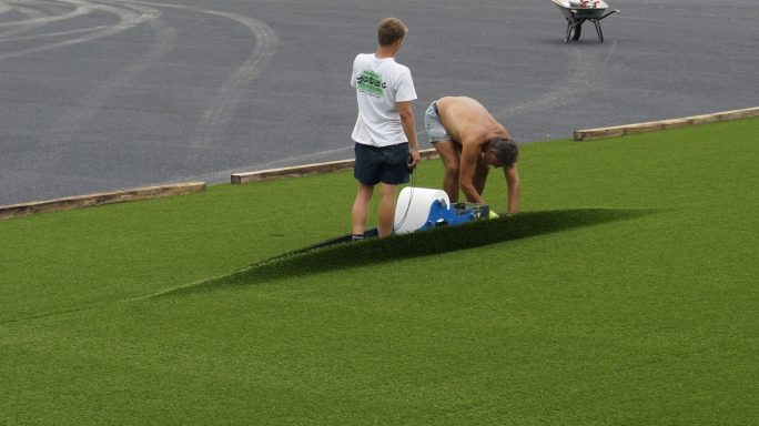 Zwei Personen arbeiten an der Verlegung von Kunstrasen auf einem Sportplatz.