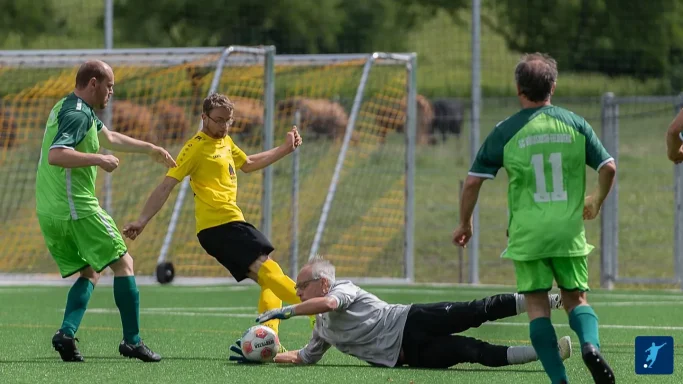 Ein Fußballspieler im gelben Trikot fällt, während ein Torwart einen Ball abwehren möchte.