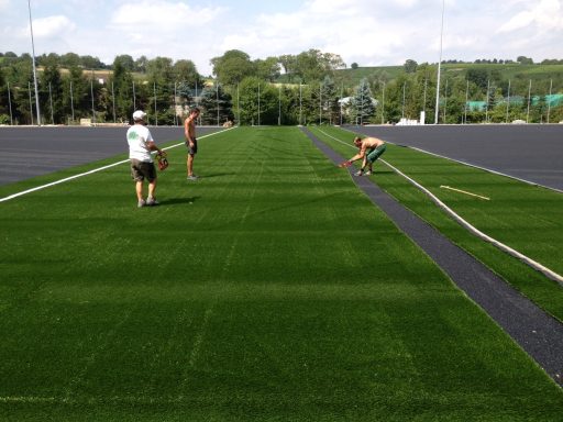 Arbeiter verlegen Kunstrasen auf einem Sportplatz bei sonnigem Wetter.